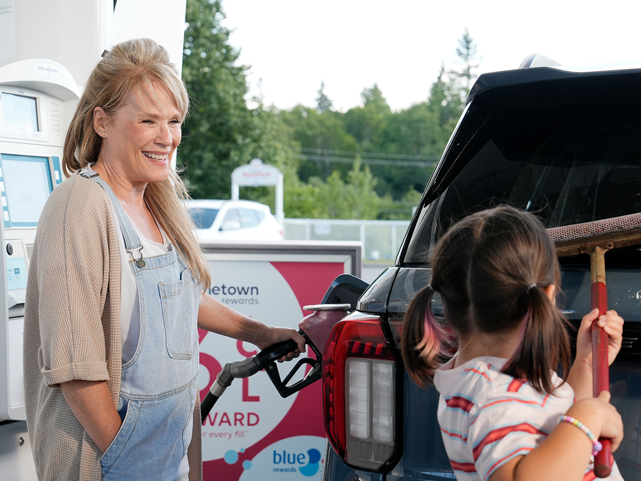 Person refuels an SUV at a gas station while another person stands beside the open cargo area holding a long-handled tool. A sign in the background advertises a “$2 reward” and “blue rewards” program.