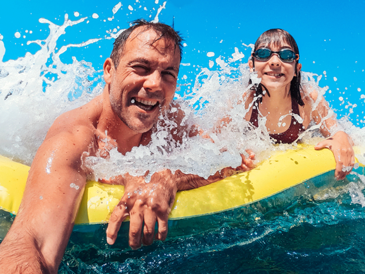 A father and daughter hold onto a yellow float in clear blue water as a wave splashes toward the camera; the daughter wears swim goggles.