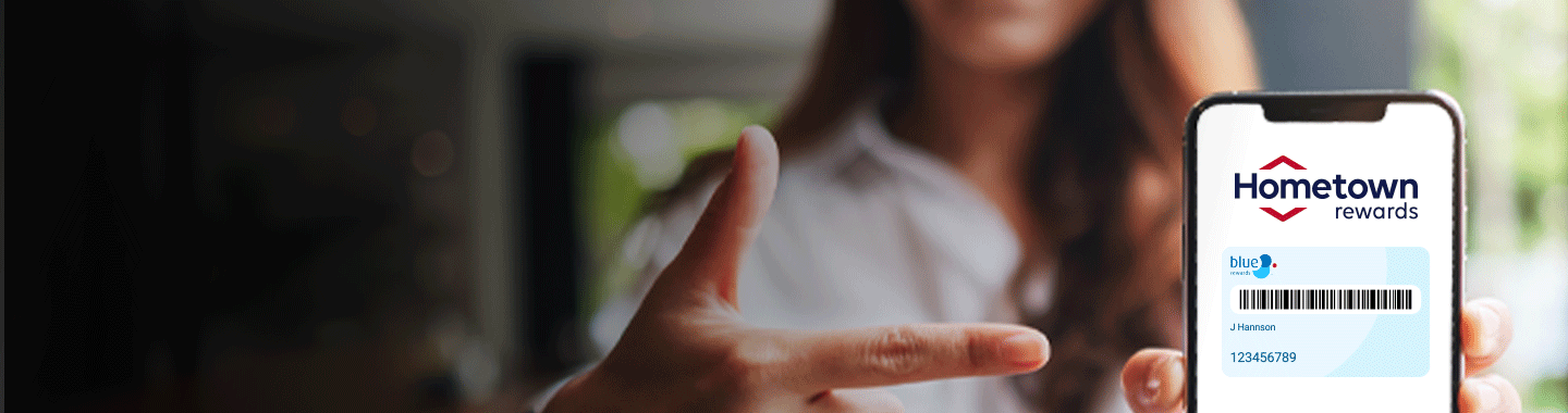 A woman with a phone in her hand displaying the Hometown Rewards app with her right hand's index finger pointing towards the phone and a thumb pointing in an upward direction.