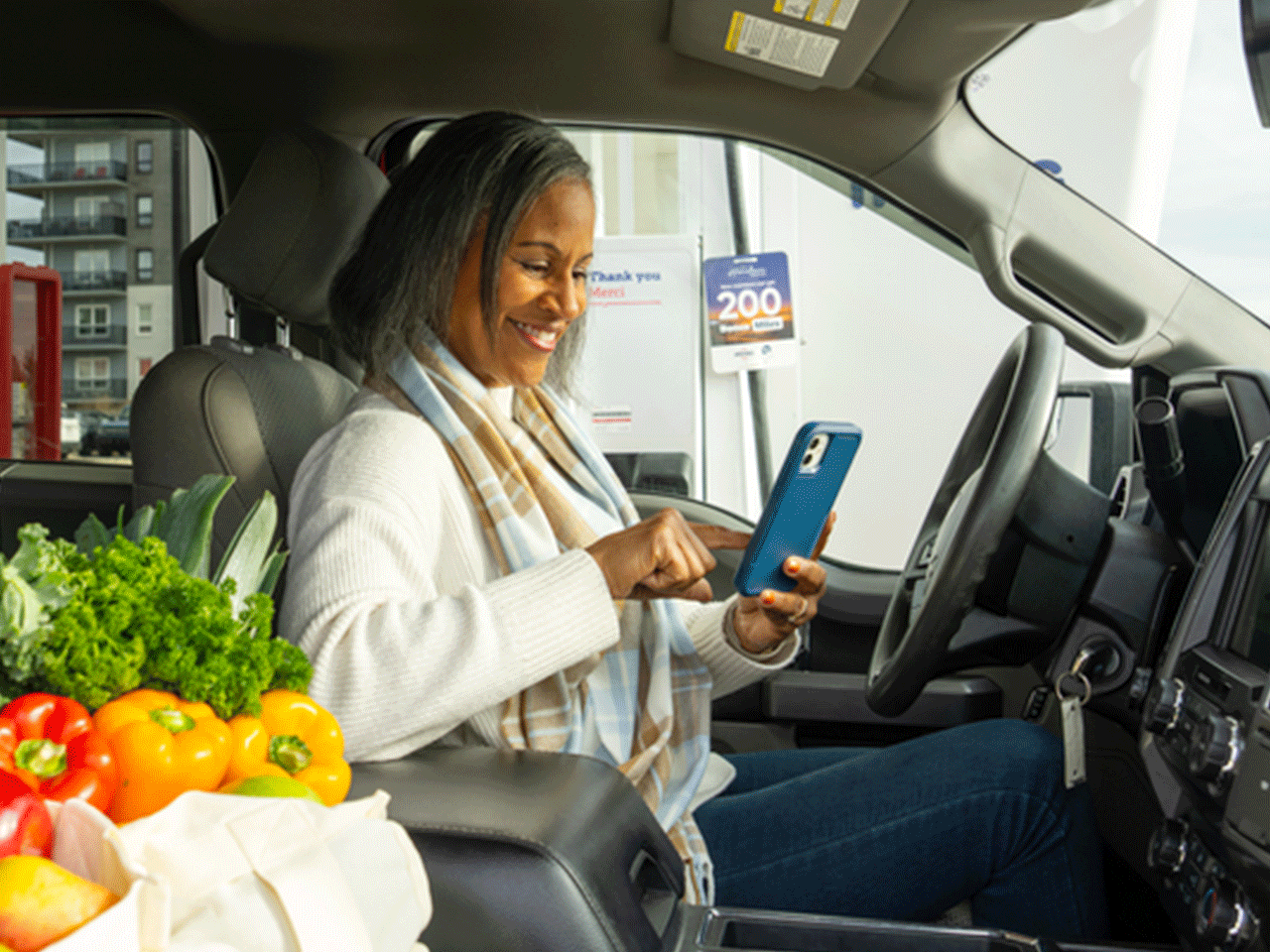 Person seated in a vehicle at an Irving gas station uses a smartphone; grocery items are visible on the passenger seat, and fuel pumps can be seen outside the window.