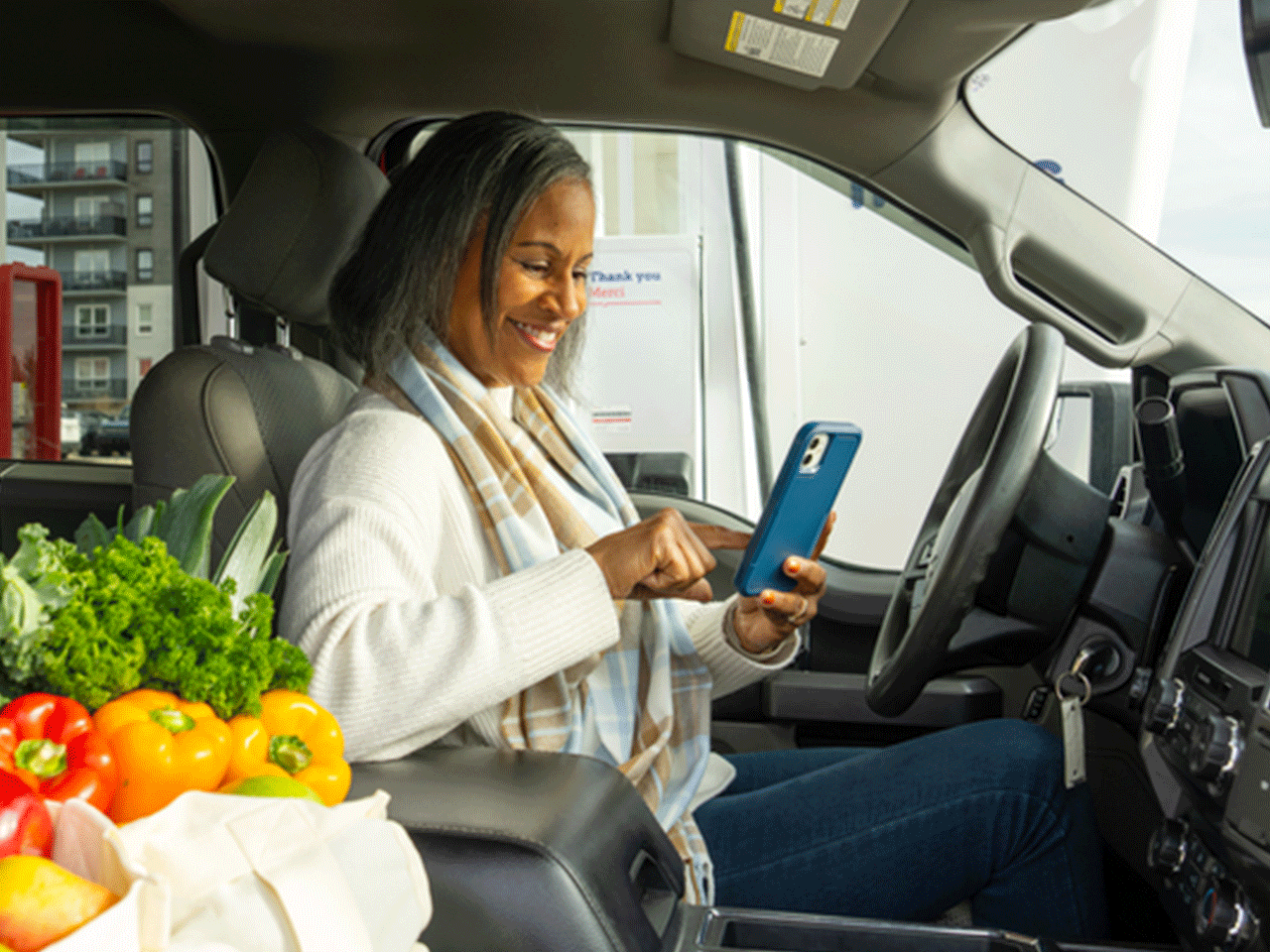 Person seated in a vehicle at a gas station taps a smartphone while groceries sit on the passenger seat; fuel pumps are visible through the window.