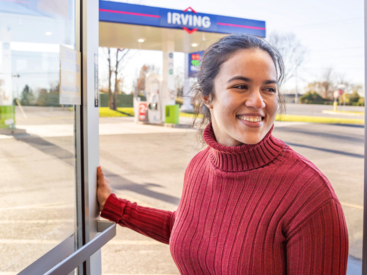 Person in a red sweater opens a glass door at an Irving gas station; fuel pumps and Irving signage are visible in the background.