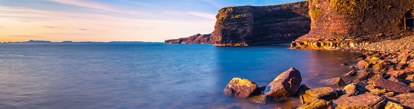 Rocky coastal cliffs at sunset overlook calm blue water, with large stones along the shoreline in the foreground and a distant horizon under a clear sky.
