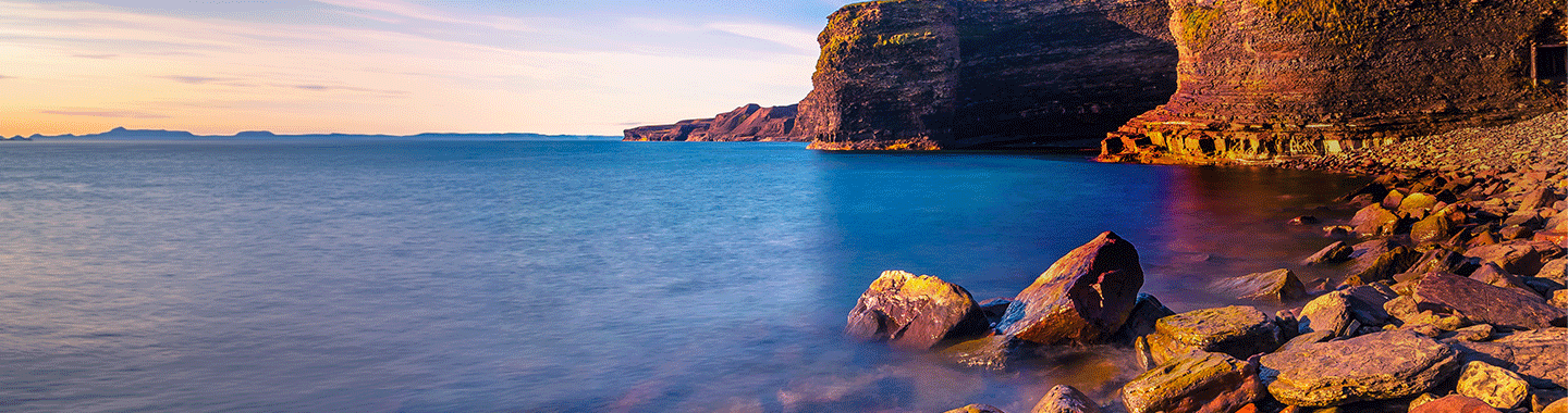 Rocky coastal cliffs at sunset above calm blue water, with large stones along the shoreline and a distant horizon under a clear sky.