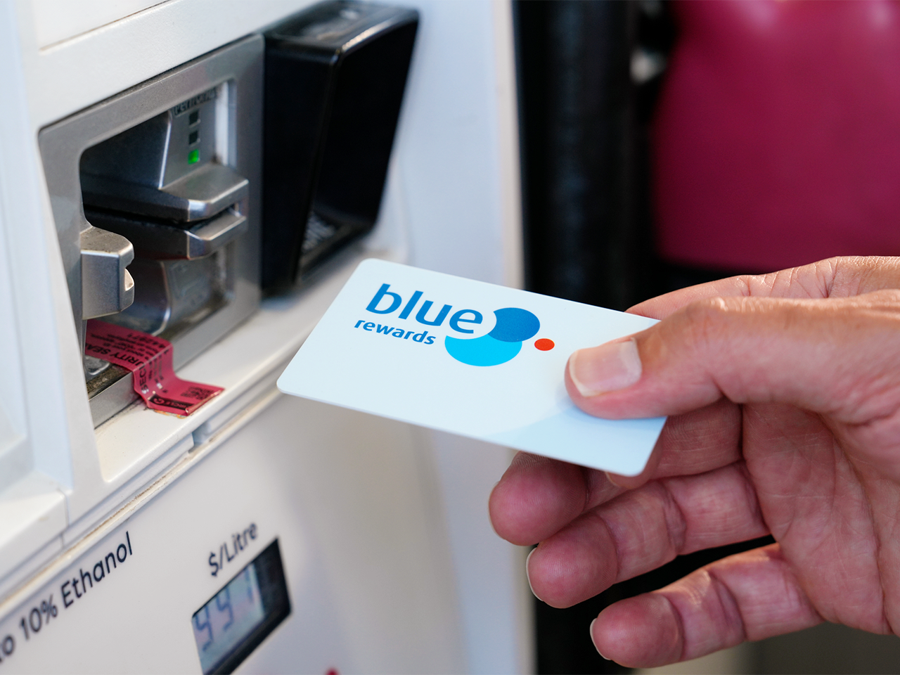 Close-up of a hand holding a Blue Rewards card near a card reader on a fuel pump. The fuel pump display and the text “Blue Rewards” are visible on the card.