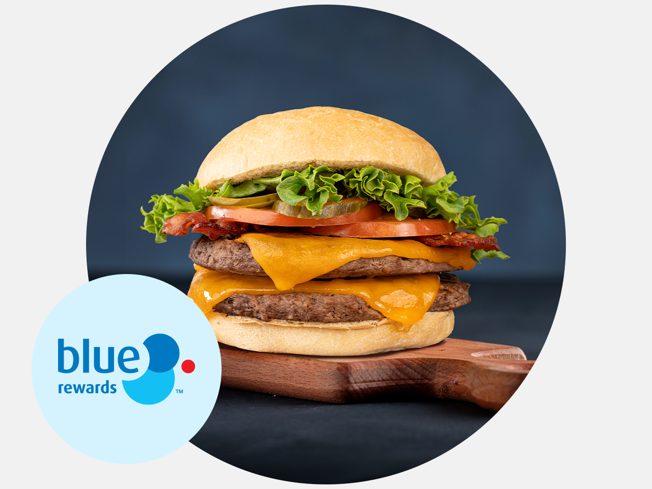 Blue Rewards logo beside a double cheeseburger on a wooden board. The burger is topped with bacon, lettuce, tomato, and pickle slices on a toasted bun.