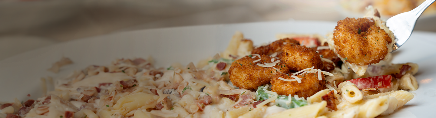 Close-up of creamy penne pasta with bacon and shredded cheese, topped with breaded shrimp. A fork lifts one shrimp above the plate.