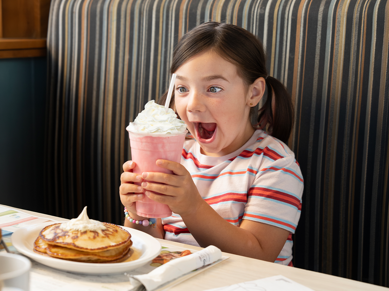Child seated in a restaurant booth holding a pink milkshake topped with whipped cream and a straw. A plate of pancakes with whipped cream is on the table in front of the child.