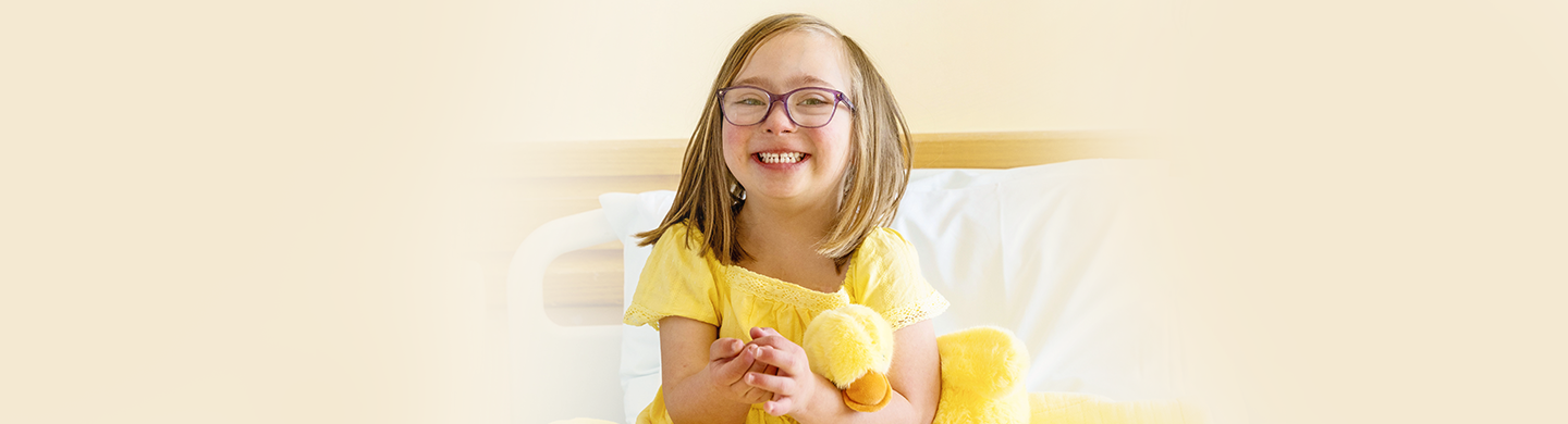 Child wearing glasses sits on a hospital bed holding a yellow plush duck, dressed in a yellow shirt against a light background.