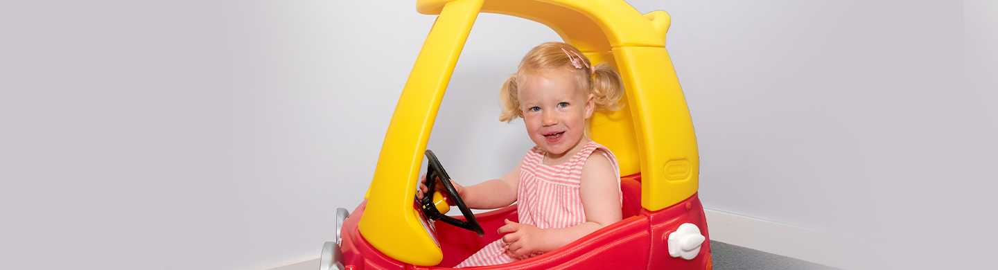 Young child sitting in a red and yellow toy car indoors, holding the steering wheel and facing the camera.