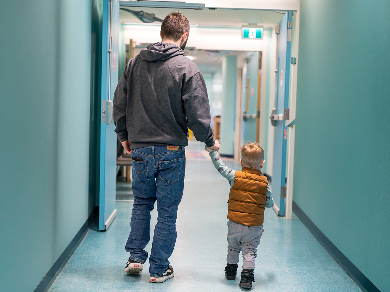An adult and a young child walk hand in hand down a brightly lit hallway with teal walls and open doors.