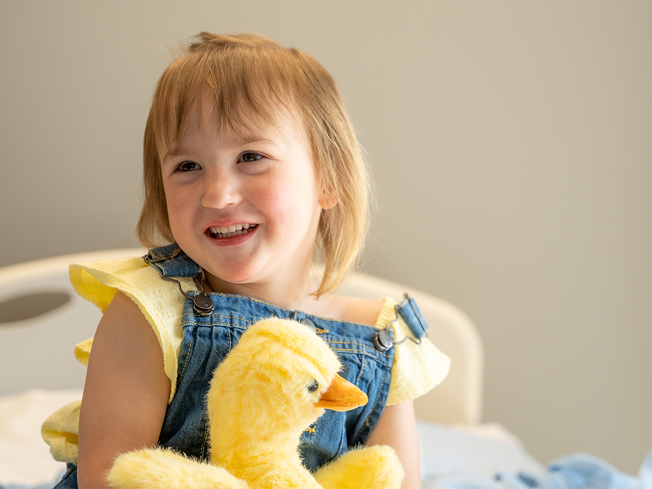 Child sitting on a hospital bed holding a yellow plush duck, wearing a yellow shirt and denim overalls.
