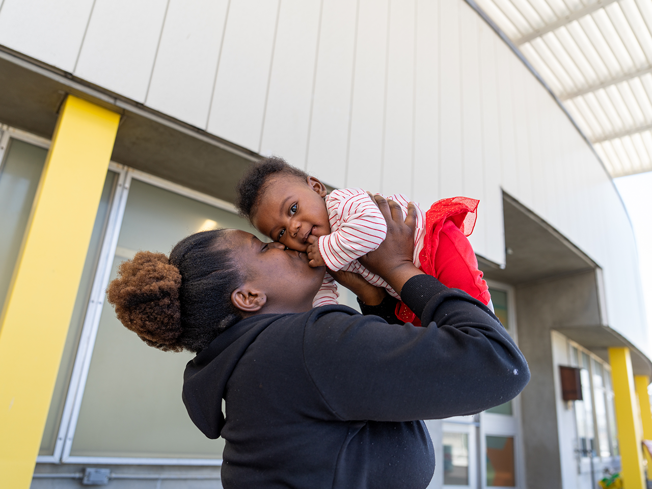 Person in a black hoodie lifts and cuddles a baby in a striped shirt and red skirt outside a modern building with yellow columns.