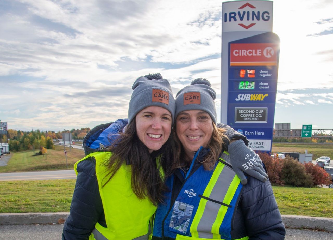 Two people wearing high-visibility safety vests and gray knit hats stand together outdoors near a roadside Irving gas station sign. The sign displays Circle K, Subway, Second Cup Coffee Co., fuel prices, and EV fast chargers. A highway, grassy area, and cloudy sky are visible in the background.