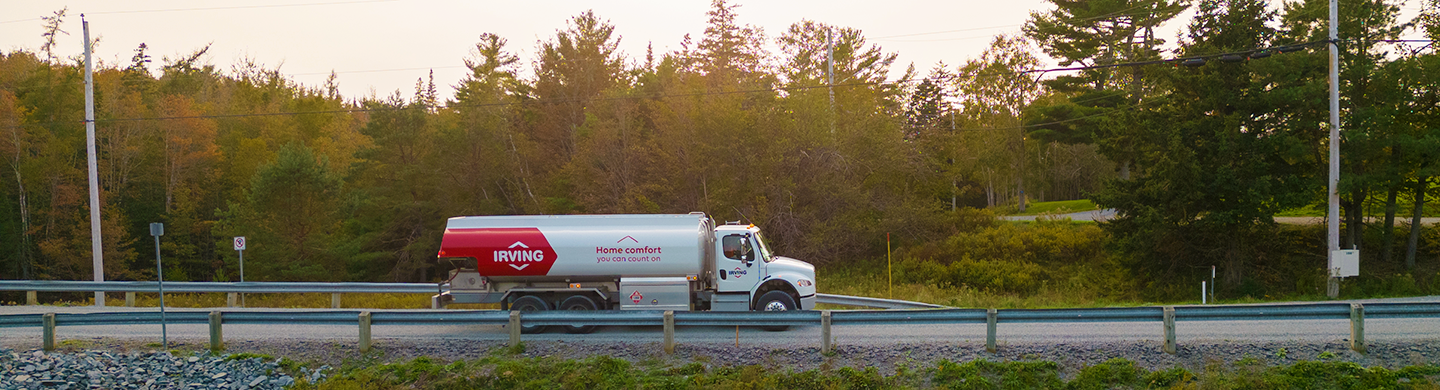 Irving fuel delivery truck driving along a rural roadside, with metal guardrails in the foreground and a wooded area of evergreen and deciduous trees in the background.