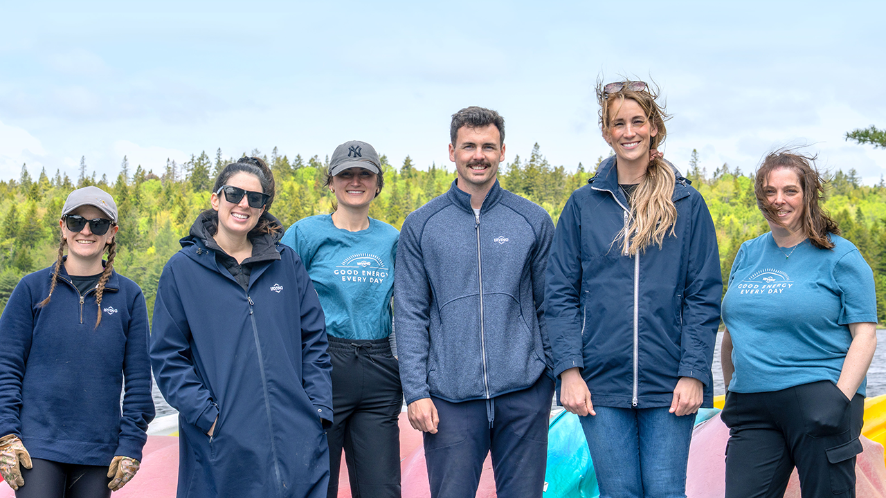 Six people standing outdoors in front of stacked canoes, with trees and water in the background. Several people wear jackets, and two wear blue shirts with the text “Good Energy Every Day.”
