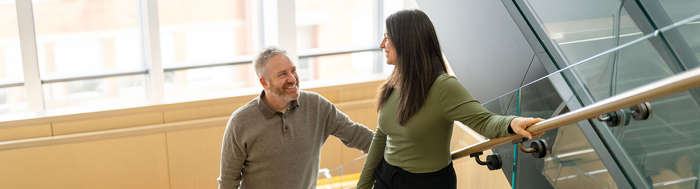 Two people on an indoor staircase in a modern office building, standing beside a glass railing and facing each other. Large windows and bright natural light fill the background.