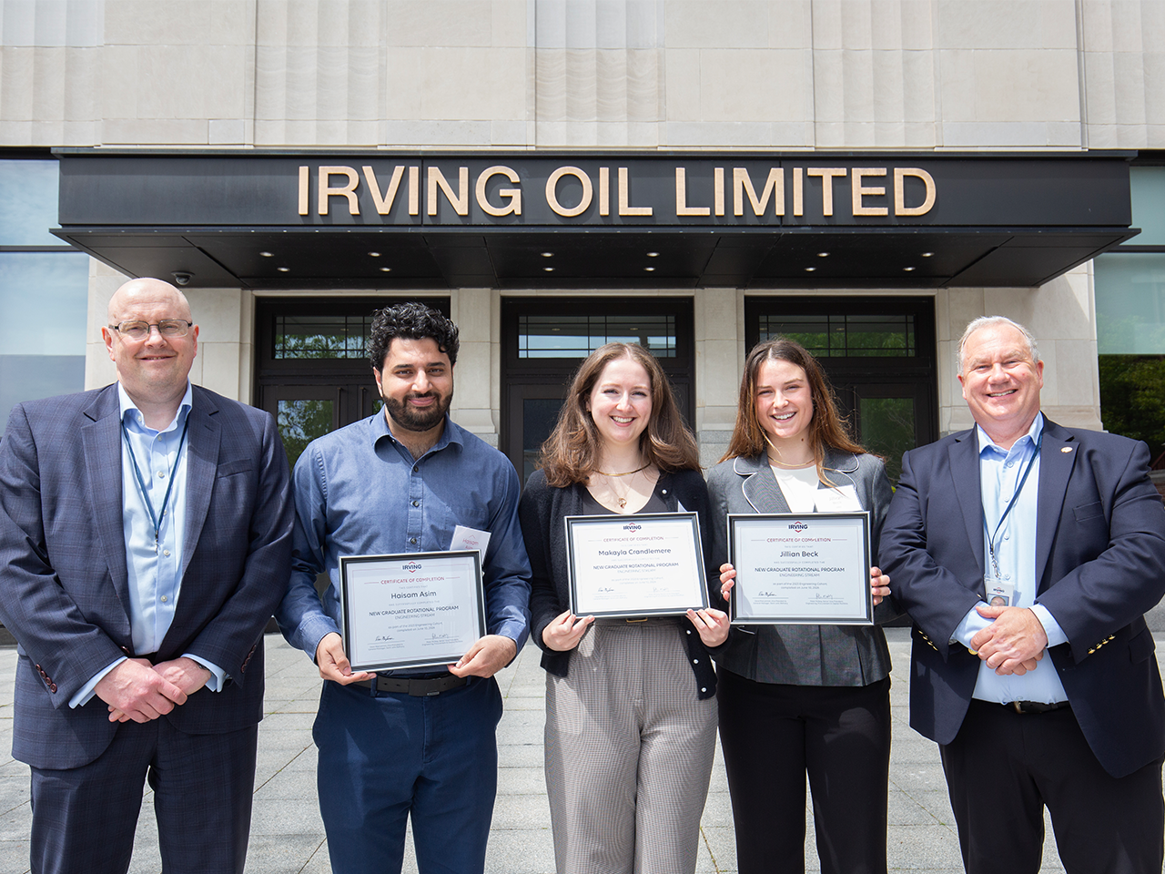Five people stand in front of the Irving Oil Limited building entrance. Three people in the center hold framed certificates, while two people in business attire stand at either side. Visible sign above the entrance reads “IRVING OIL LIMITED.”