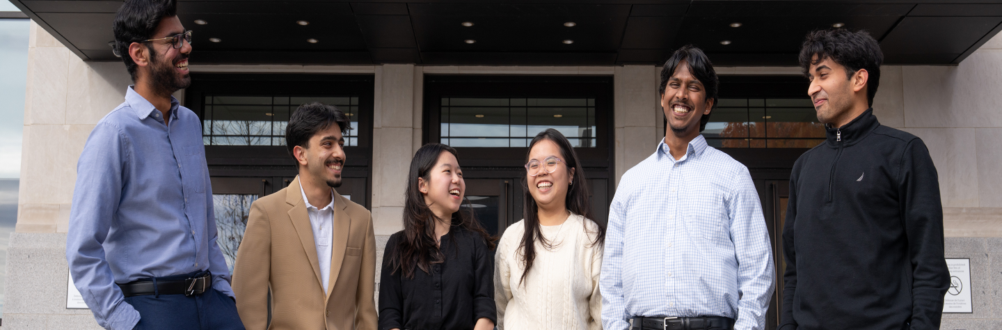 Six people stand together outside a building entrance, facing one another in conversation. The group wears a mix of business-casual and casual clothing, with glass doors and stone walls visible in the background.