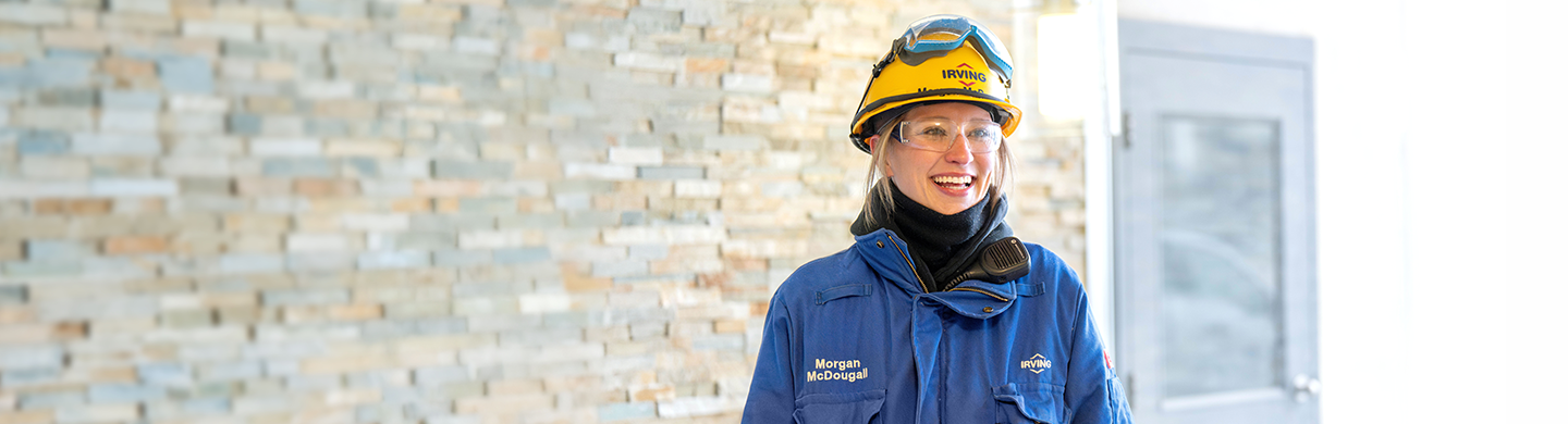 Refinery facility technician Morgan McDougall poses in PPE with refinery in background.