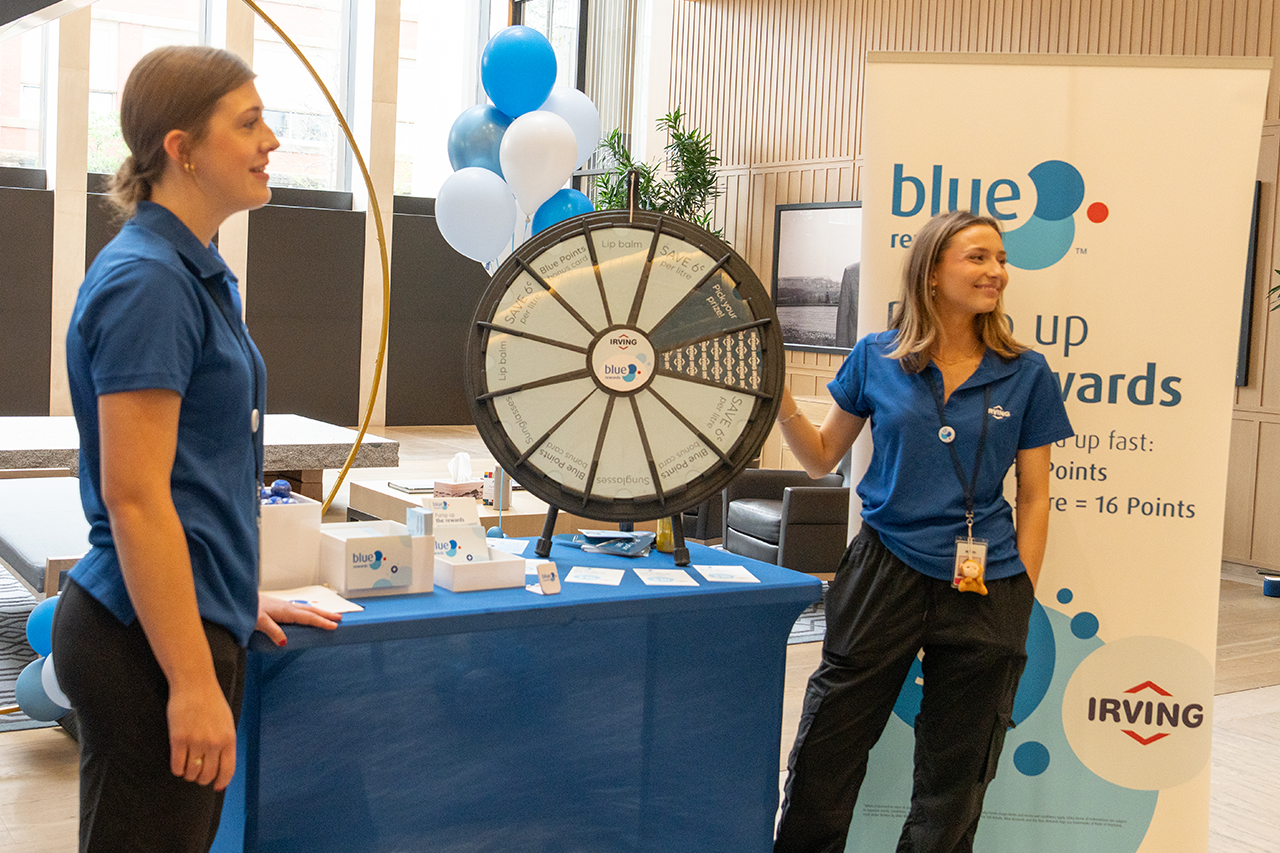 Two people stand at a promotional booth with a prize wheel. A banner beside the display reads “blue rewards” and includes the Irving logo, while balloons and giveaway items are arranged on a table.