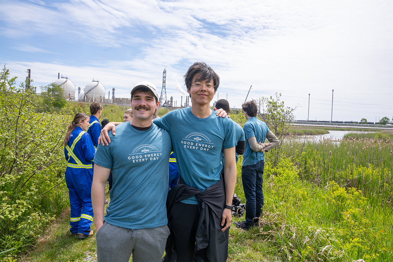 Two people wearing “Good Energy Every Day” T‑shirts stand together in a grassy wetland area, with several other volunteers nearby and industrial facilities visible in the background.