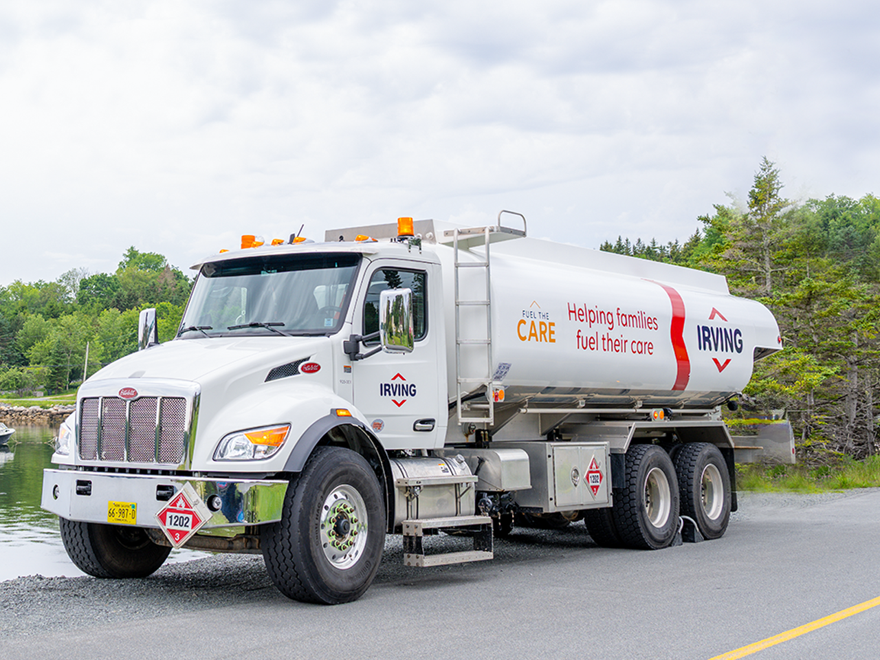 White Irving fuel delivery truck parked on a roadside beside a body of water and trees. The tanker displays the text “Fuel the Care” and “Helping families fuel their core,” along with Irving branding. Hazard placards are visible on the front and side of the vehicle under a cloudy sky.