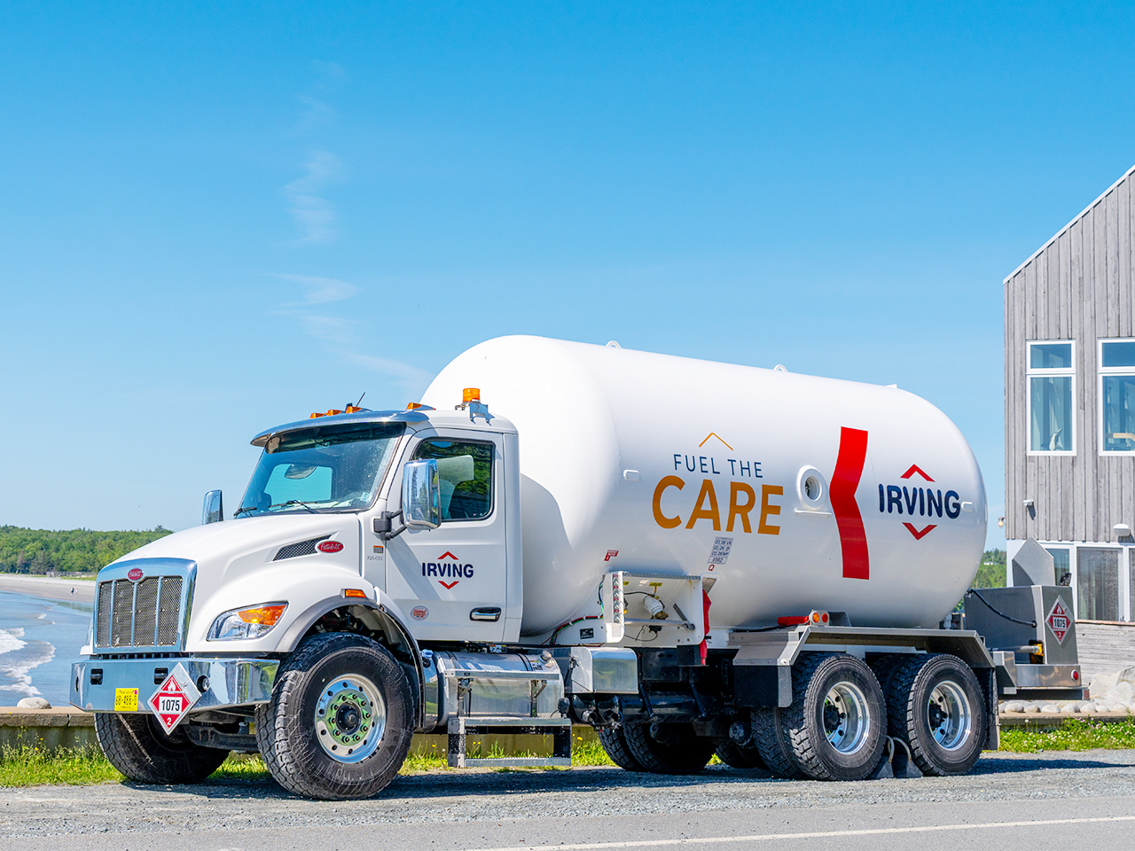 White Irving propane delivery truck parked beside a coastal roadway near a building and shoreline. The tank displays the text “Fuel the Care” and Irving branding. Hazard placards are visible on the front and rear of the vehicle, with a clear blue sky and water in the background.