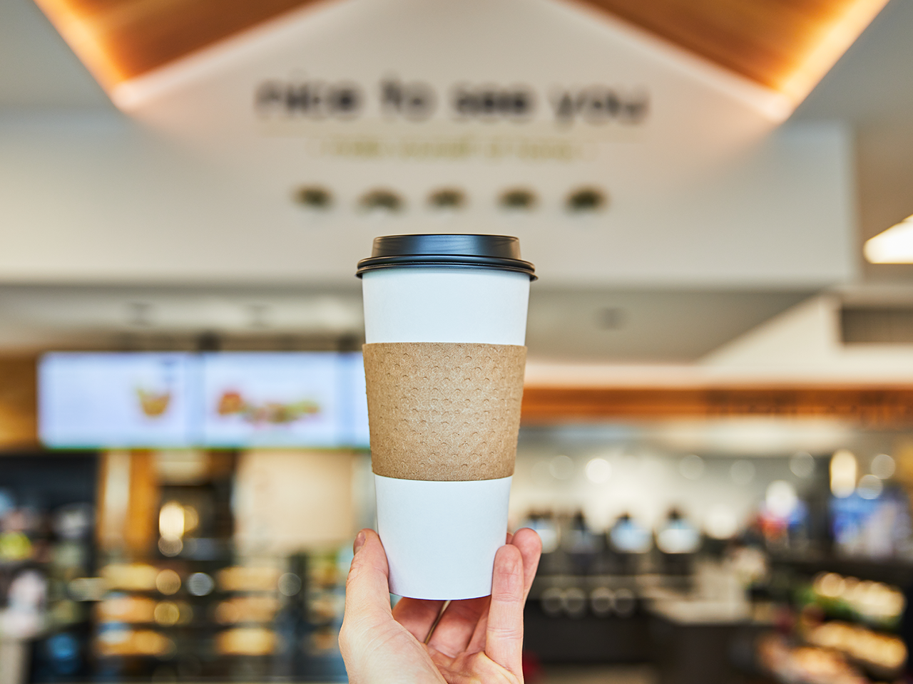 Hand holding a white takeaway coffee cup with a black lid and recycled cardboard sleeve in the foreground, centered against a softly blurred coffee shop interior with menu screens and a sign reading “Pick up coffee for you” in the background.