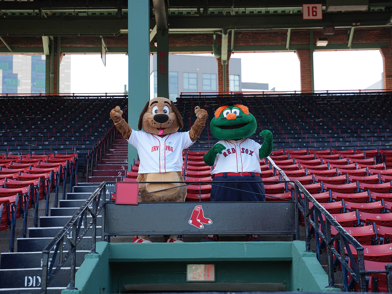 Two baseball mascots wearing Red Sox jerseys stand in a seating section at Fenway Park, raising their arms among rows of red stadium seats. A Red Sox logo is visible on the railing in front of them.