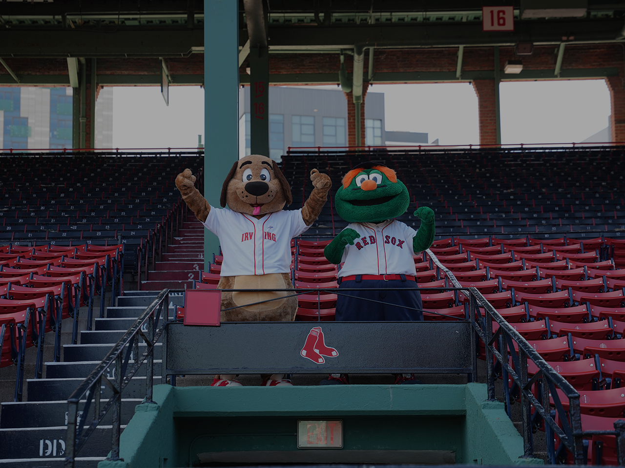 Two baseball team mascots wearing Red Sox jerseys stand in a seating section at Fenway Park, with rows of red stadium seats and a Red Sox logo visible in the foreground.
