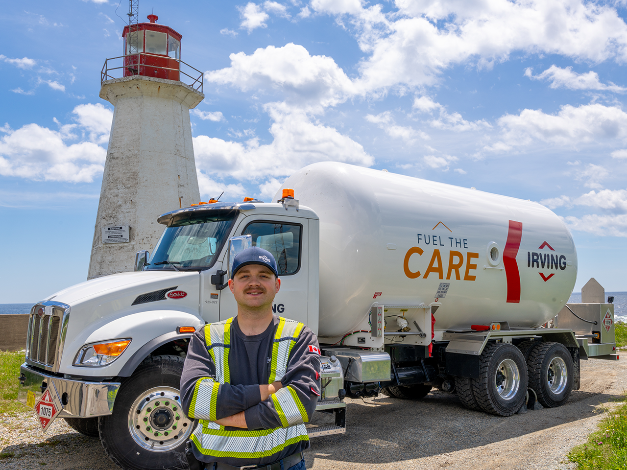 Person wearing a high-visibility safety jacket and hardhat-style cap stands with arms crossed in front of a white Irving fuel tanker truck parked beside a white lighthouse with a red lantern room. The tanker displays the text “FUEL THE CARE” and “IRVING.” Blue sky with scattered clouds in the background.