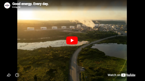 Aerial view of an Irving Oil refinery beside wetlands and a winding road at sunrise; storage tanks and industrial buildings emit steam. Overlaid text reads “Good energy. Every day. Irving Oil,” with a YouTube play button centered.