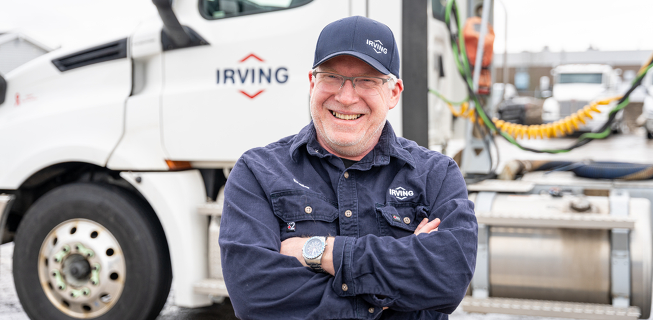 Person in an Irving work uniform and cap standing with arms crossed in front of an Irving transport truck, with the truck cab and equipment visible in the background.